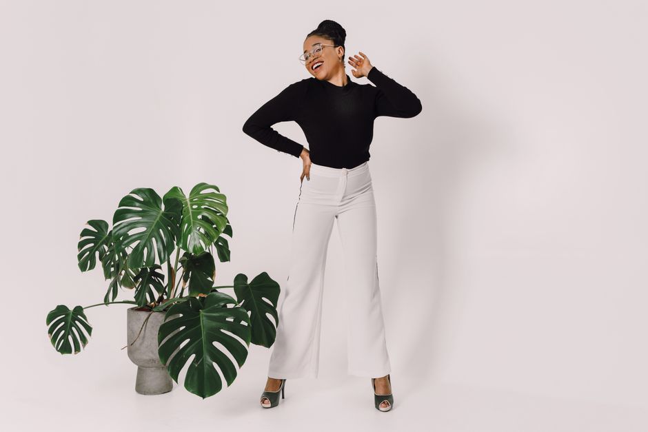 Fashionable woman in black and white attire posing next to Monstera plant on a white background.