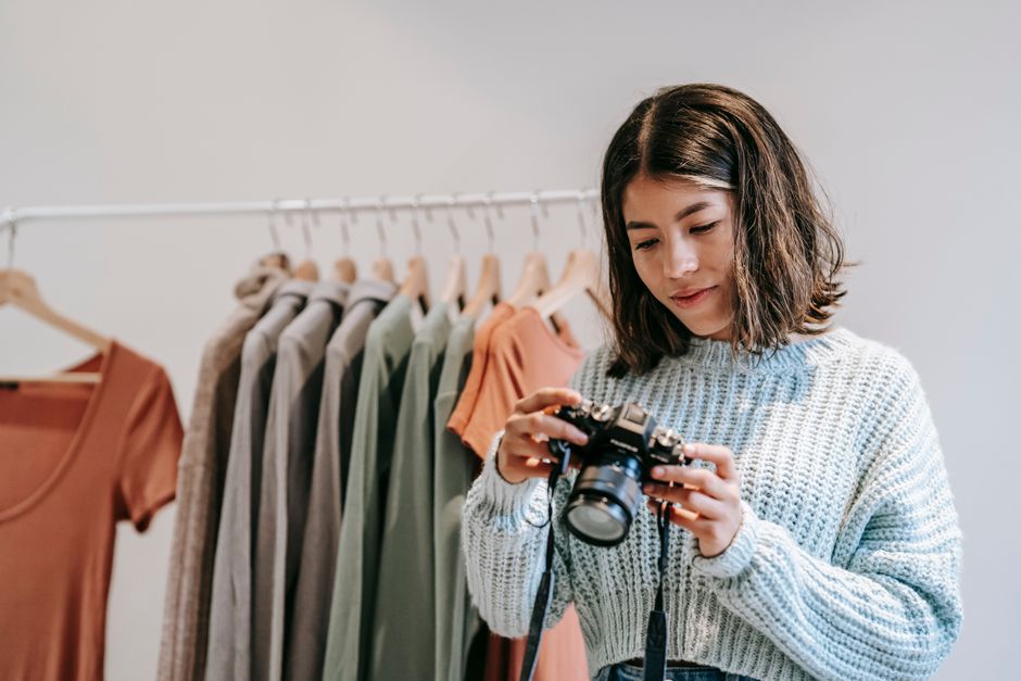 Young Latin American woman in knitted sweater watching professional photo camera against stand with different apparel on light background