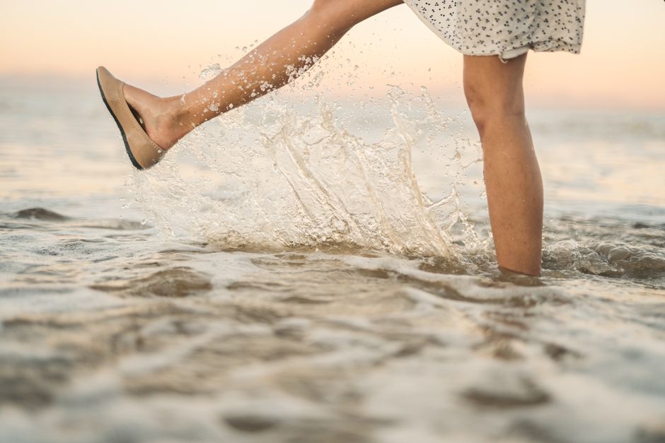 A woman in a floral dress kicks ocean water, creating a splash at sunset.