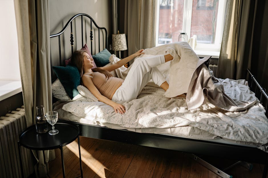 Pregnant woman comfortably lying in bed, enjoying morning sunlight in cozy bedroom setting.