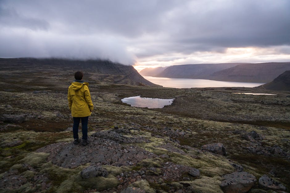 Back view of faceless traveler in yellow raincoat standing on rocky surface in front of sea between ridge covered with mist under cloudy sky at sunset