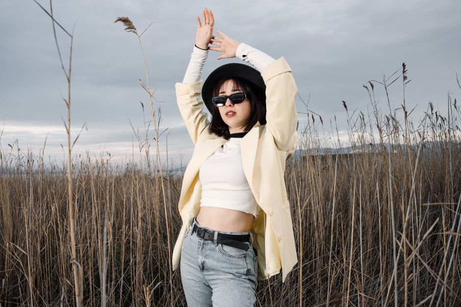 Woman wearing a yellow coat and sunglasses poses in a field of reeds under a cloudy sky.