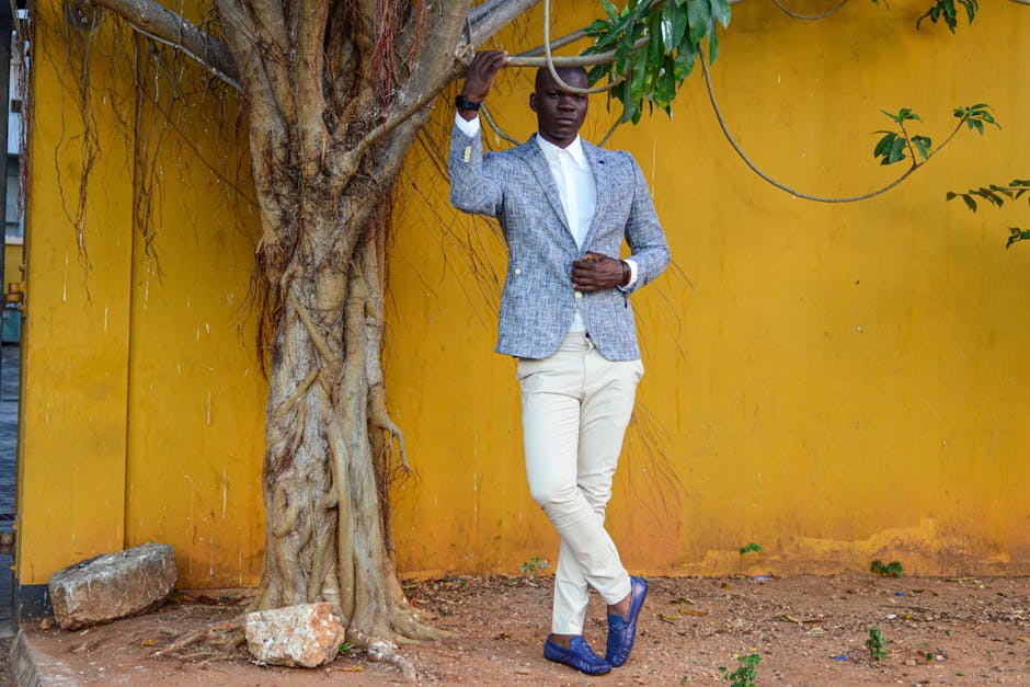 Man in a stylish outfit posing against a vibrant yellow wall beside a tree.