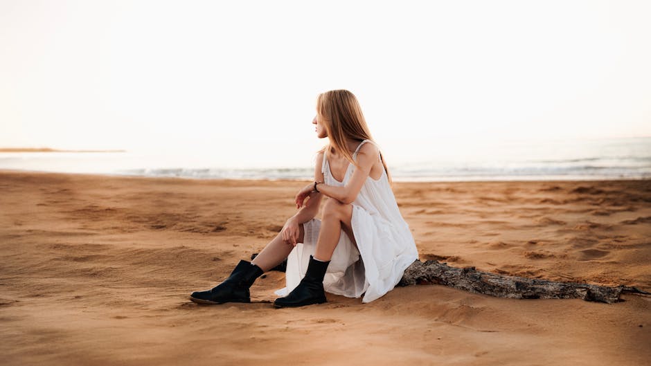 Woman in white dress sitting on a peaceful sandy beach during the day.