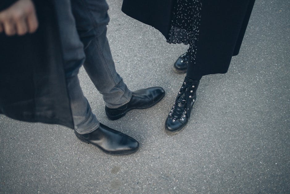 A fashionable couple shows off their black leather footwear while standing together on a city street.