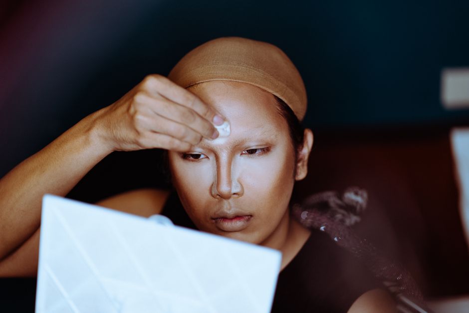 Concentrated ethnic female applying powder and rouge on face on blurred background of room