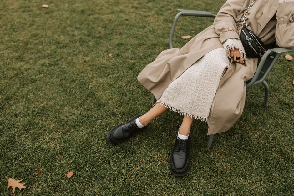Stylish person in trench coat sitting on metal chair with grass background.