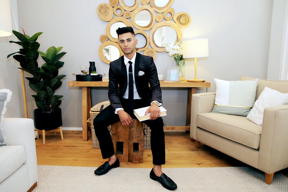 A fashionable young man in a suit sitting in a stylish, modern living room with wooden decor.