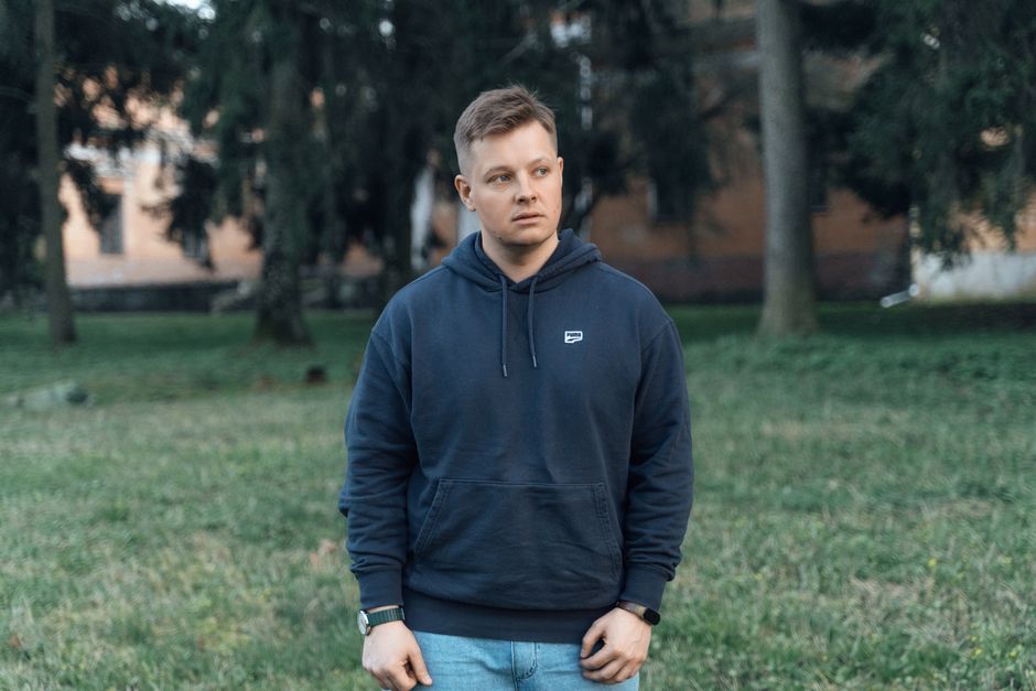 A casual portrait of a man in a hoodie standing outdoors in a grassy park setting.