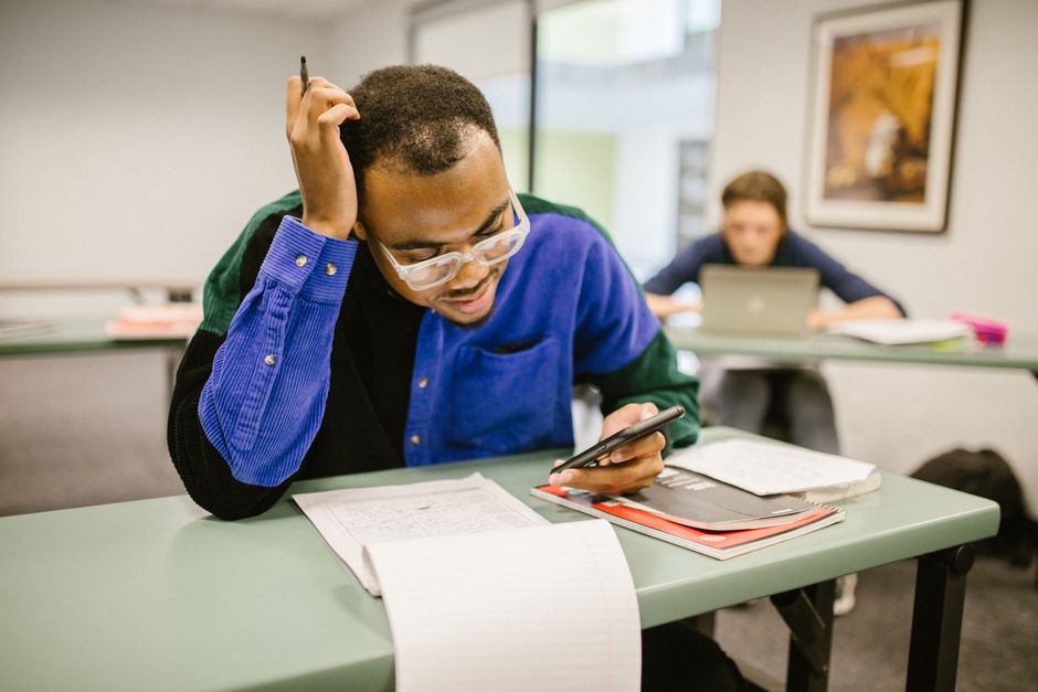 A college student studying in class using a smartphone, with papers and books on the desk.