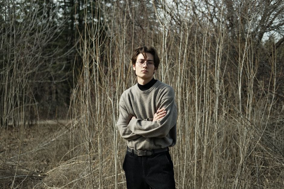 A fashionable man posing amidst tall, dry plants in a rural setting.