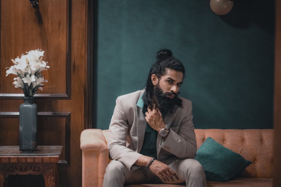 A fashionable man with a beard sits thoughtfully on a couch indoors.