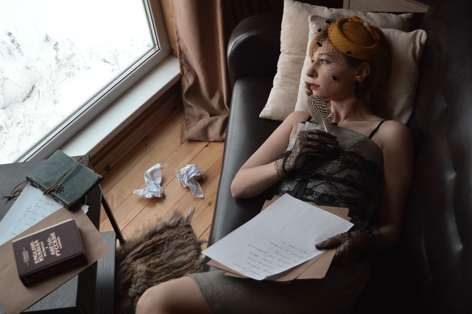 High angle of young wistful female author in vintage wear with feather looking away on couch against window