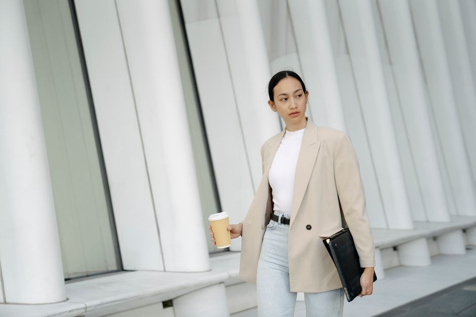 Confident Asian female office worker in formal clothes carrying takeaway coffee and folder while looking away with attention