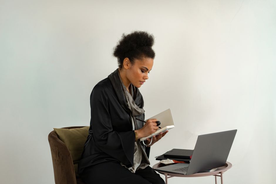 Woman in black dress taking notes while working remotely with a laptop indoors.