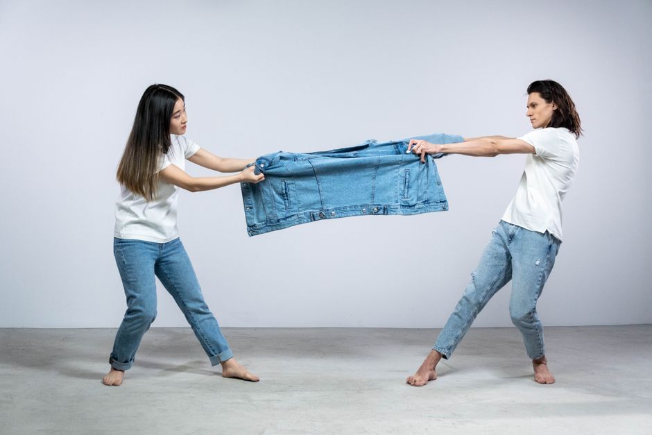 Two adults pulling on a denim jacket in a playful fashion pose in a studio setting.