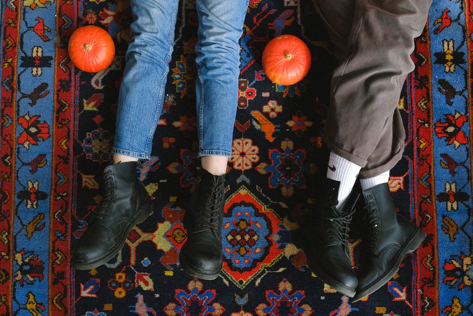 Two pairs of legs with leather boots lie on a vibrant carpet, flanked by pumpkins.