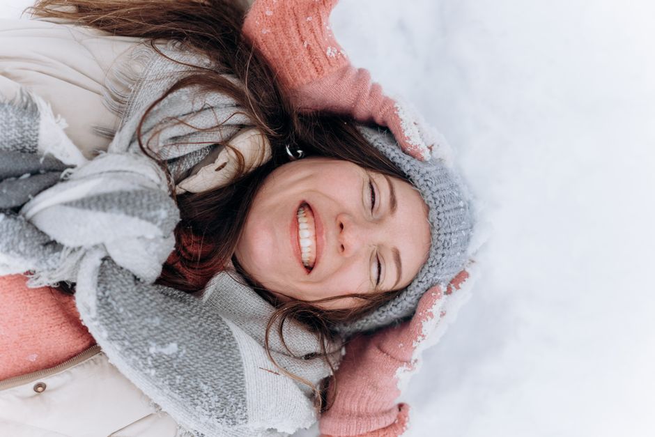 Smiling woman in winter attire enjoying the snow while lying down outdoors.