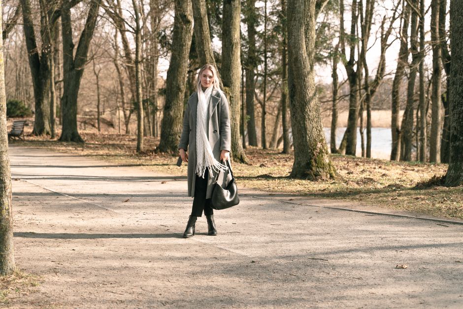 A woman in a gray coat walks through a forest park path on a clear spring day.