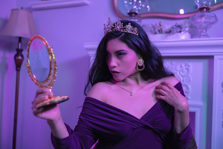 A woman wearing a crown and elegant attire gazes into a handheld mirror indoors.