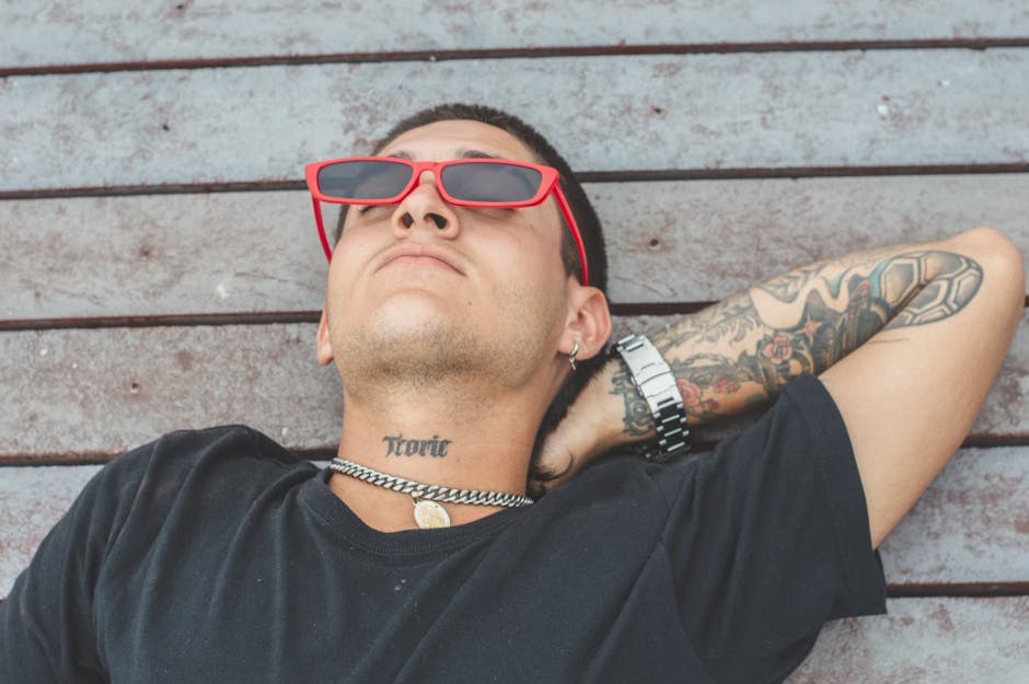 A young man in sunglasses relaxes outdoors, showcasing tattoos and casual style.