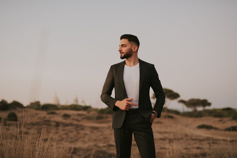 A fashionable man in a suit poses outdoors in a serene desert-like landscape at sunset.