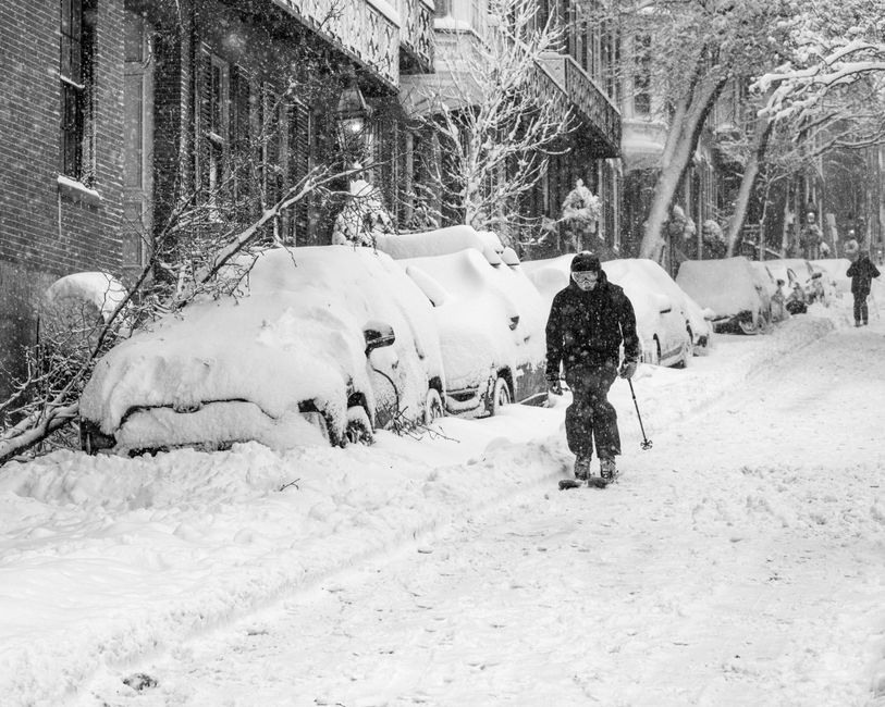 A skier navigates a snowy Boston street, flanked by snow-covered cars and trees, during a heavy snowfall.