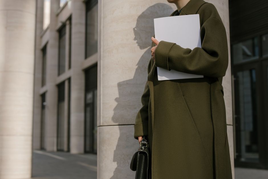 Woman in a green coat holding documents outside a modern office building.