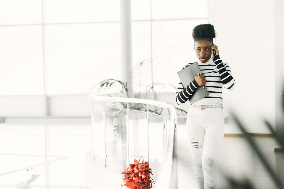 Young African American businesswoman talking on her phone while holding a laptop indoors.